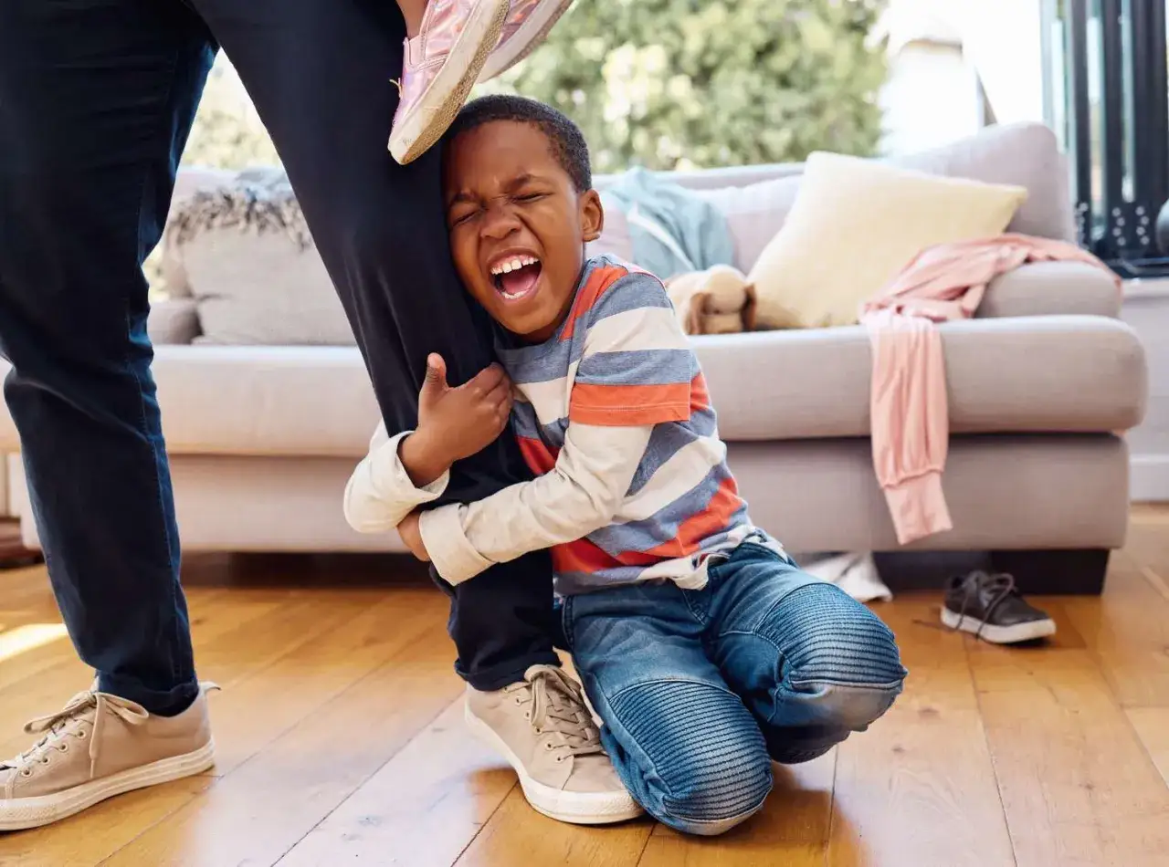 Ni&ntilde;o llora y abraza la pierna de su padre, mostrando un apego excesivo. En el sof&aacute;, un peluche y ropa.