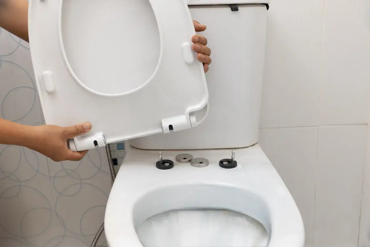 Montaż deski WC. Dłonie trzymają białą deskę, przygotowując ją do zamocowania na sedesie.