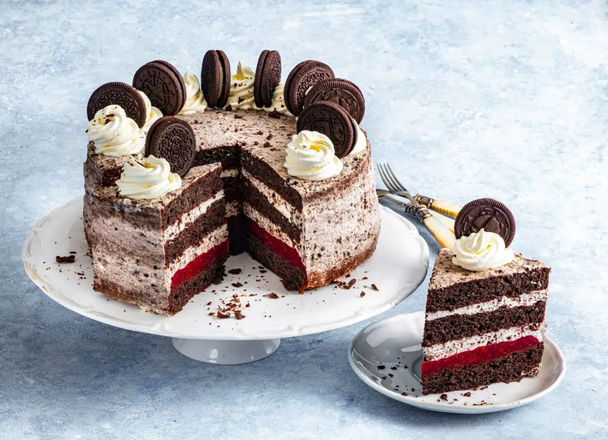 Przepis na tort oreo: czekoladowe blaty przełożone kremem i malinową galaretką, ozdobiony ciasteczkami oreo i bitą śmietaną.