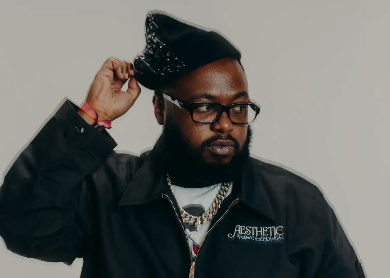 A man in a black beanie and jacket, with a diamond chain, adjusts his hat. He's known for his contributions to the James Fauntleroy discography.