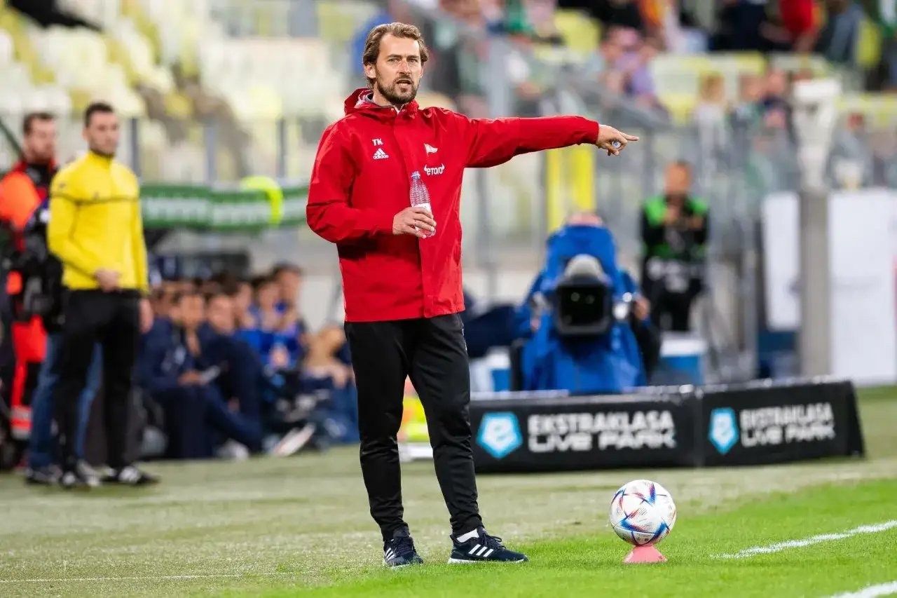 Tomasz Kaczmarek, trener w czerwonej kurtce, wskazuje palcem na boisko, trzymając butelkę wody.