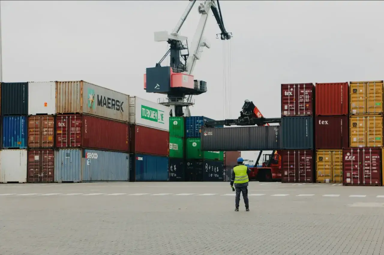 Ein Arbeiter &uuml;berwacht den Umschlag von Containern, w&auml;hrend ein Kran und ein Gabelstapler im Einsatz sind. Dies ist ein typischer Vorgang f&uuml;r die **import customs clearance**.