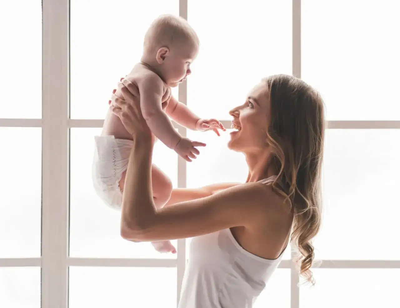 Szczęśliwa mama podnosi do g&oacute;ry swoje 7 miesięczne dziecko, kt&oacute;re w pieluszce radośnie wyciąga rączki.