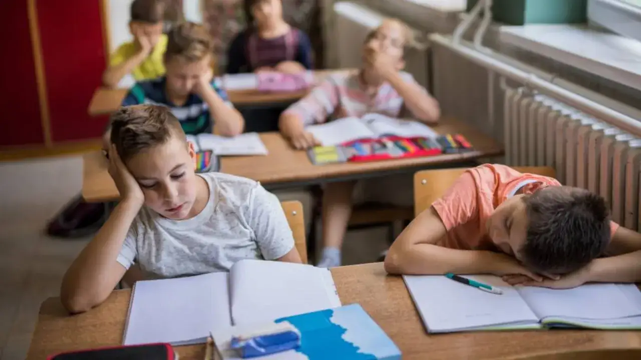 Ni&ntilde;os aburridos en un aula, reflejo de la educaci&oacute;n tradicional. Uno apoya la cabeza en la mano, otro duerme sobre su libro.