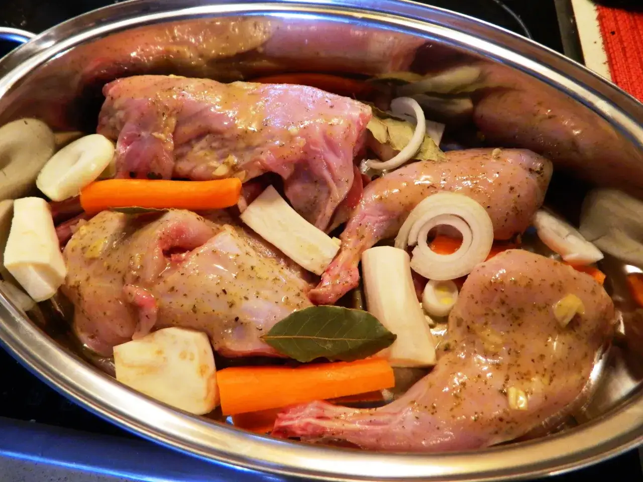 Kawałki królika w naczyniu, gotowe do pieczenia. Marynata do królika pieczonego w piekarniku zawiera marchew, pietruszkę, cebulę i liście laurowe.