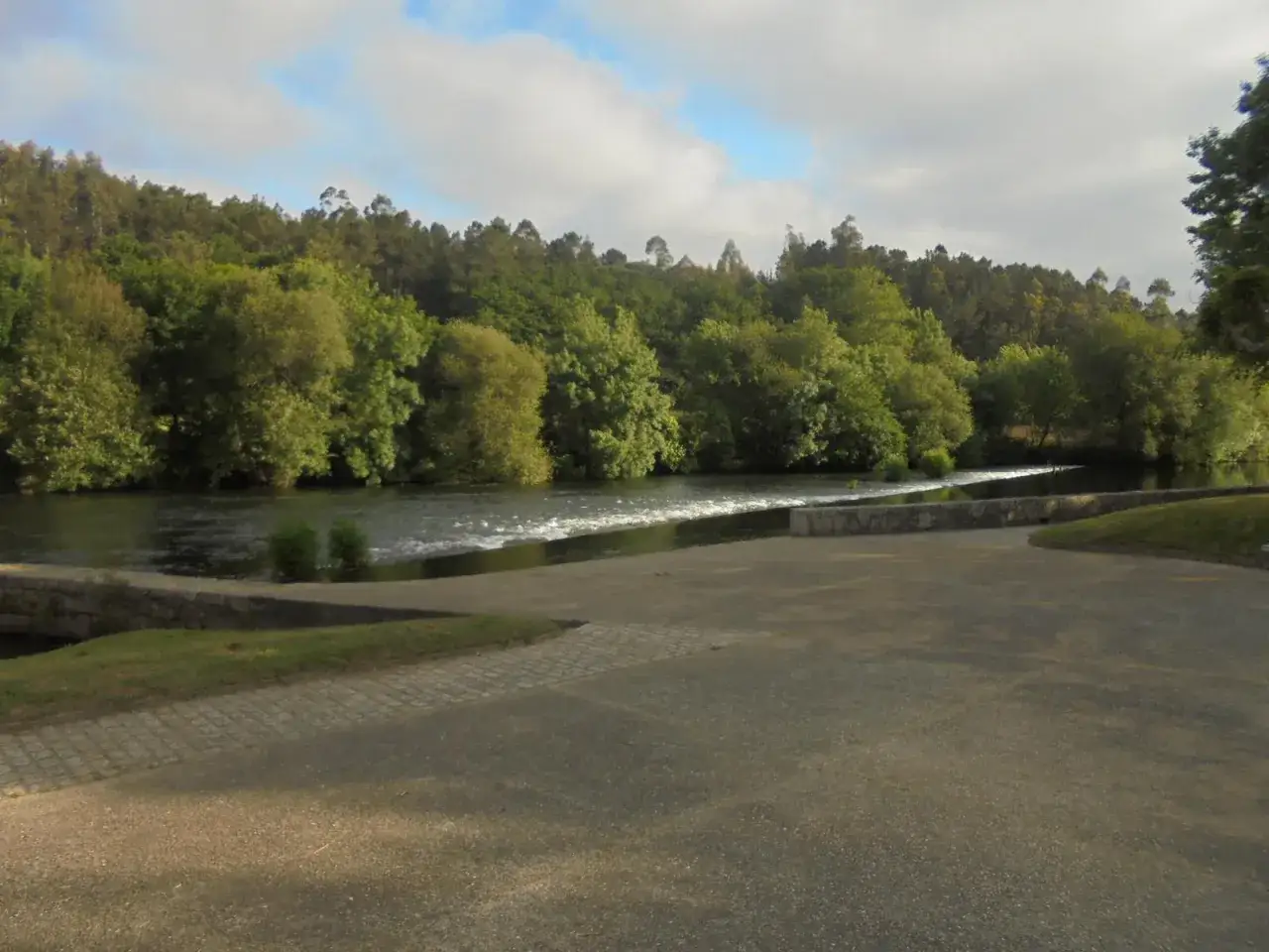 Un d&iacute;a soleado en la playa fluvial Maceira, con el r&iacute;o fluyendo entre &aacute;rboles frondosos y un cielo nublado.