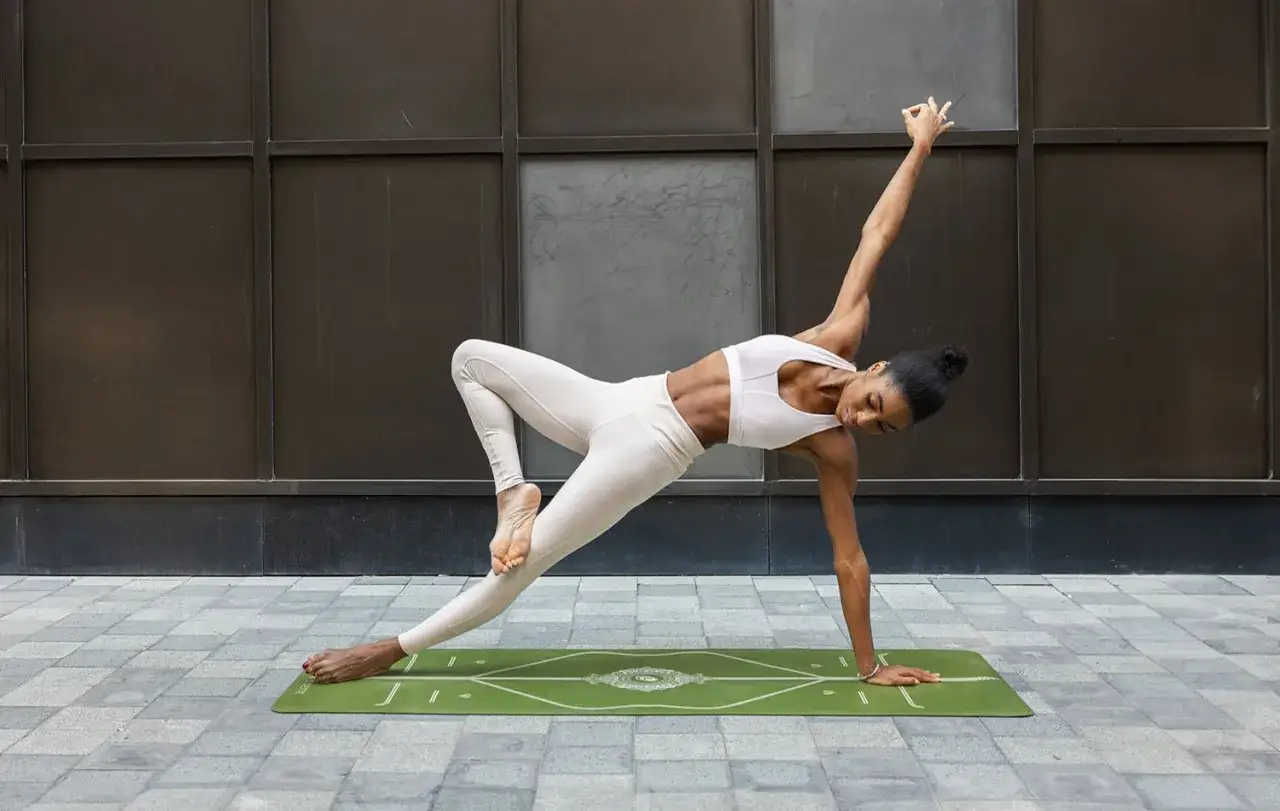 Frau in beiger Sportkleidung übt eine herausfordernde Yoga-Pose auf einer grünen Matte. Ihre linke Hand stützt sie, während ihr rechtes Bein über das linke geschwungen ist.