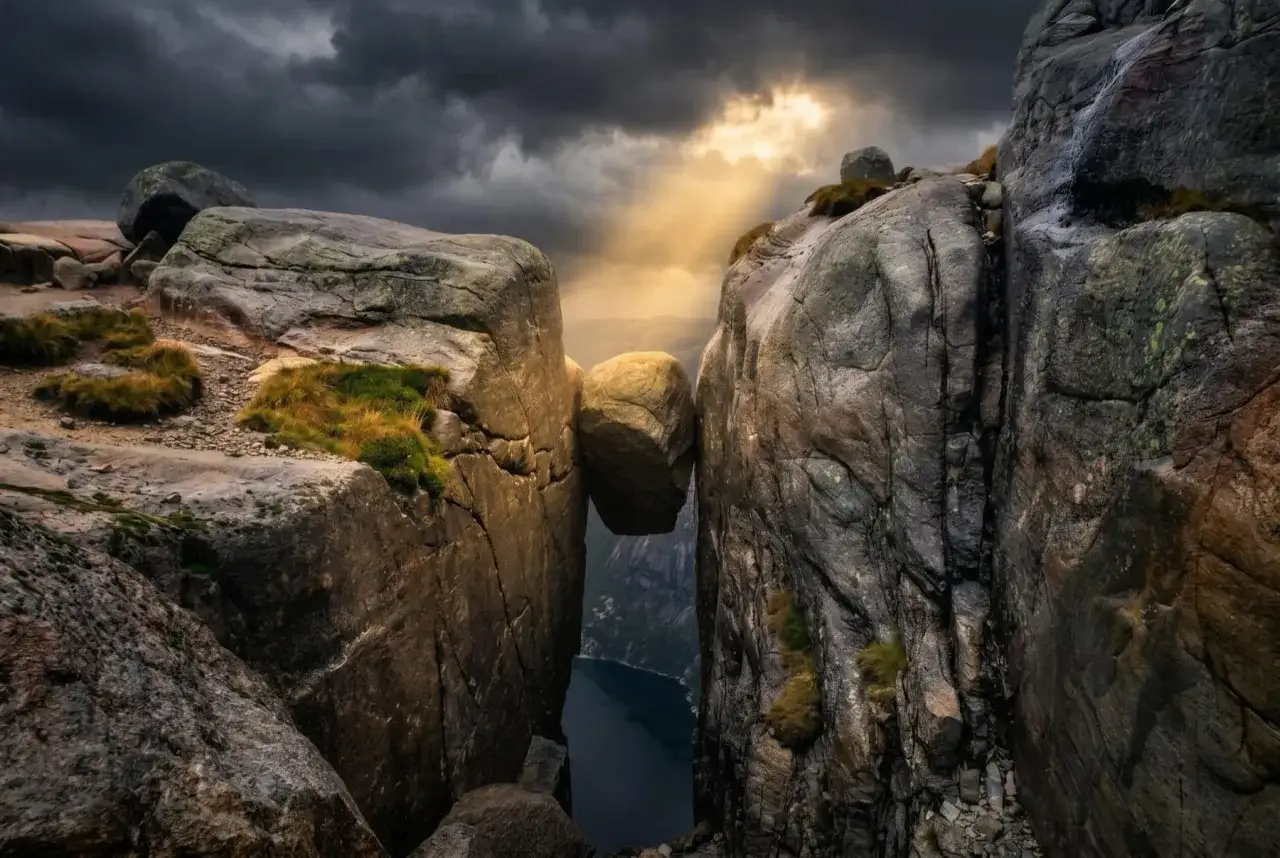 Enorm sten balanserar mellan klippor i Norge. Dramatiskt ljus bryter igenom molnen &ouml;ver en djup fjord.