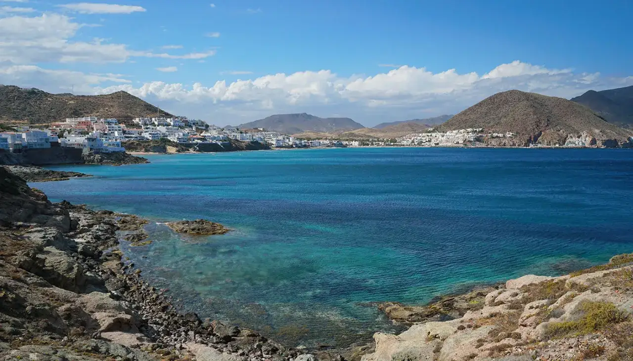 T&uuml;rkisblaues Meer vor einer K&uuml;stenstadt mit wei&szlig;en H&auml;usern, die sich an H&uuml;gel schmiegen. Ein malerischer Huelva Strand mit felsigem Ufer.