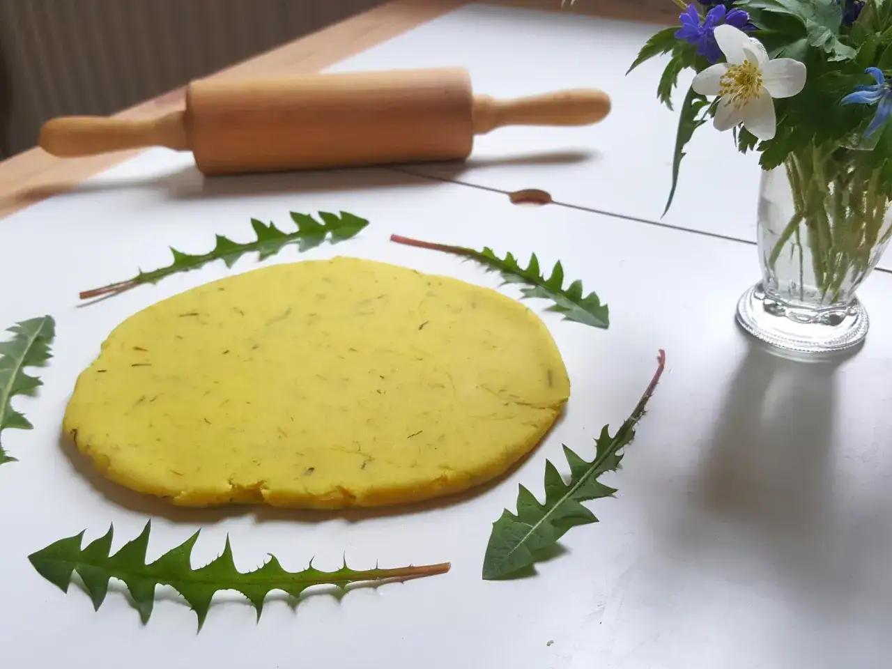 Gul deg, kavel och maskrosblad ligger redo f&ouml;r ett maskros recept. En vas med vilda blommor st&aring;r i bakgrunden.
