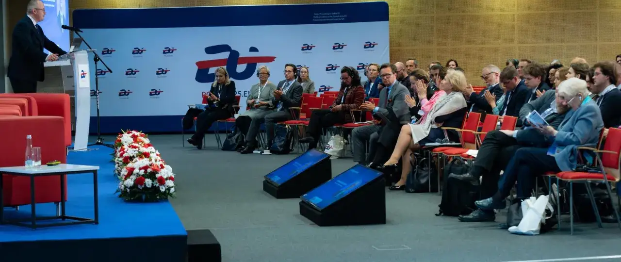 Ludzie na sali konferencyjnej słuchają przemówienia. W tle logo z napisem 