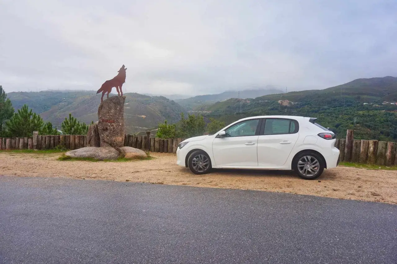 Ein wei&szlig;er Kleinwagen parkt neben einer Wolfsstatue auf einem Felsen mit Blick auf eine h&uuml;gelige Landschaft. Tolle Carjet Erfahrungen auf dieser Reise!