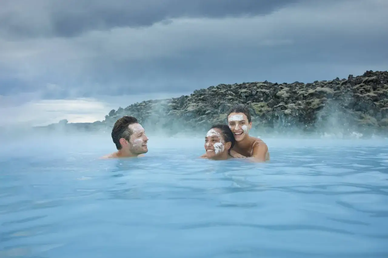 Trzy uśmiechnięte osoby z białymi maskami na twarzach kąpią się w turkusowej wodzie. W tle widać parę i skaliste wzgórza. To relaks w blue lagoon.
