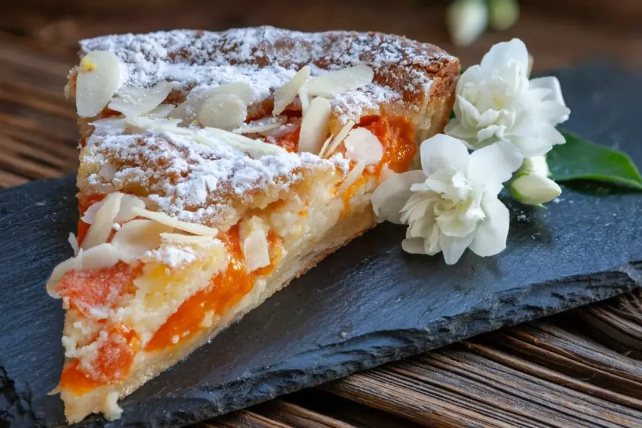 Ein Stück weltbester Aprikosenkuchen, bestäubt mit Puderzucker und Mandelblättchen, daneben zarte weiße Blüten.