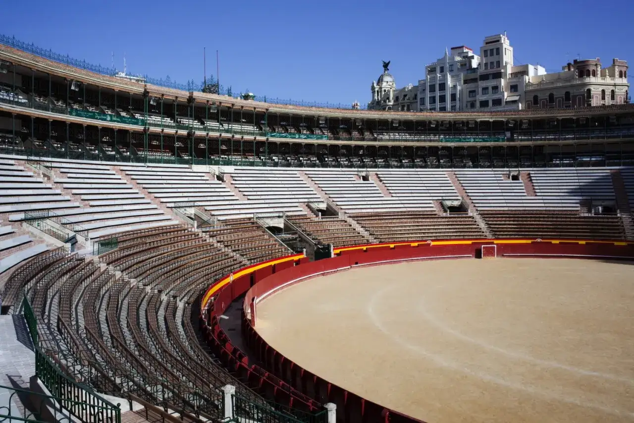 Interior de una plaza de toros vacía, con la arena de arena y la contrabarrera roja. Las gradas de madera y piedra se curvan alrededor.
