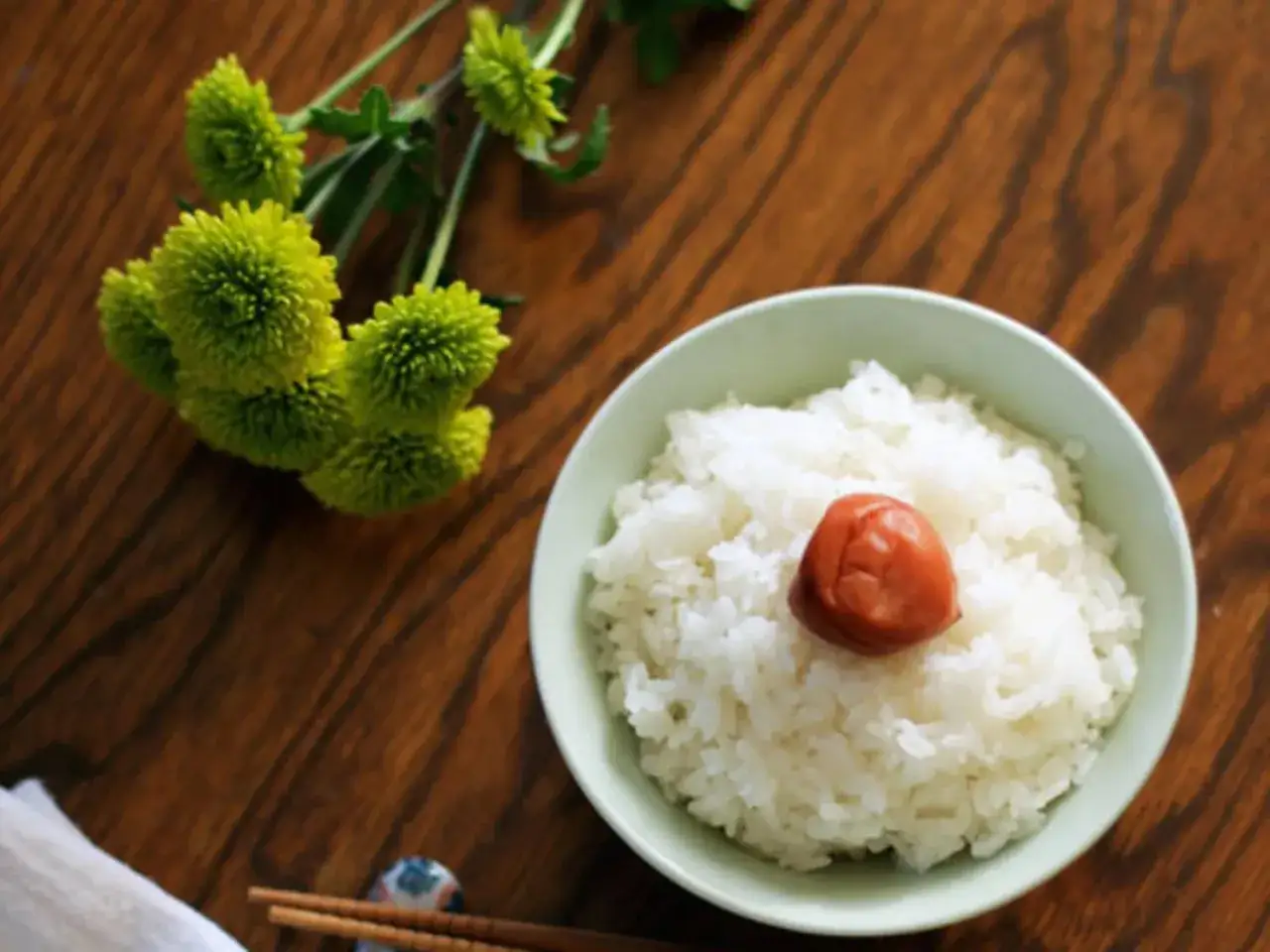 A bowl of perfectly cooked Japanese rice, topped with a pickled plum, sits on a wooden table next to green flowers. This image inspires learning how to cook Japanese rice.