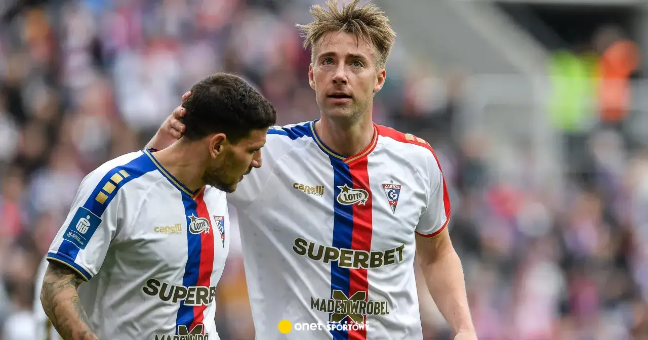 Dwóch piłkarzy Górnika Zabrze w białych strojach z czerwono-niebieskimi pasami. Jeden z nich, z blond włosami, patrzy przed siebie, drugi, z ciemnymi włosami, ma dłoń na jego ramieniu.