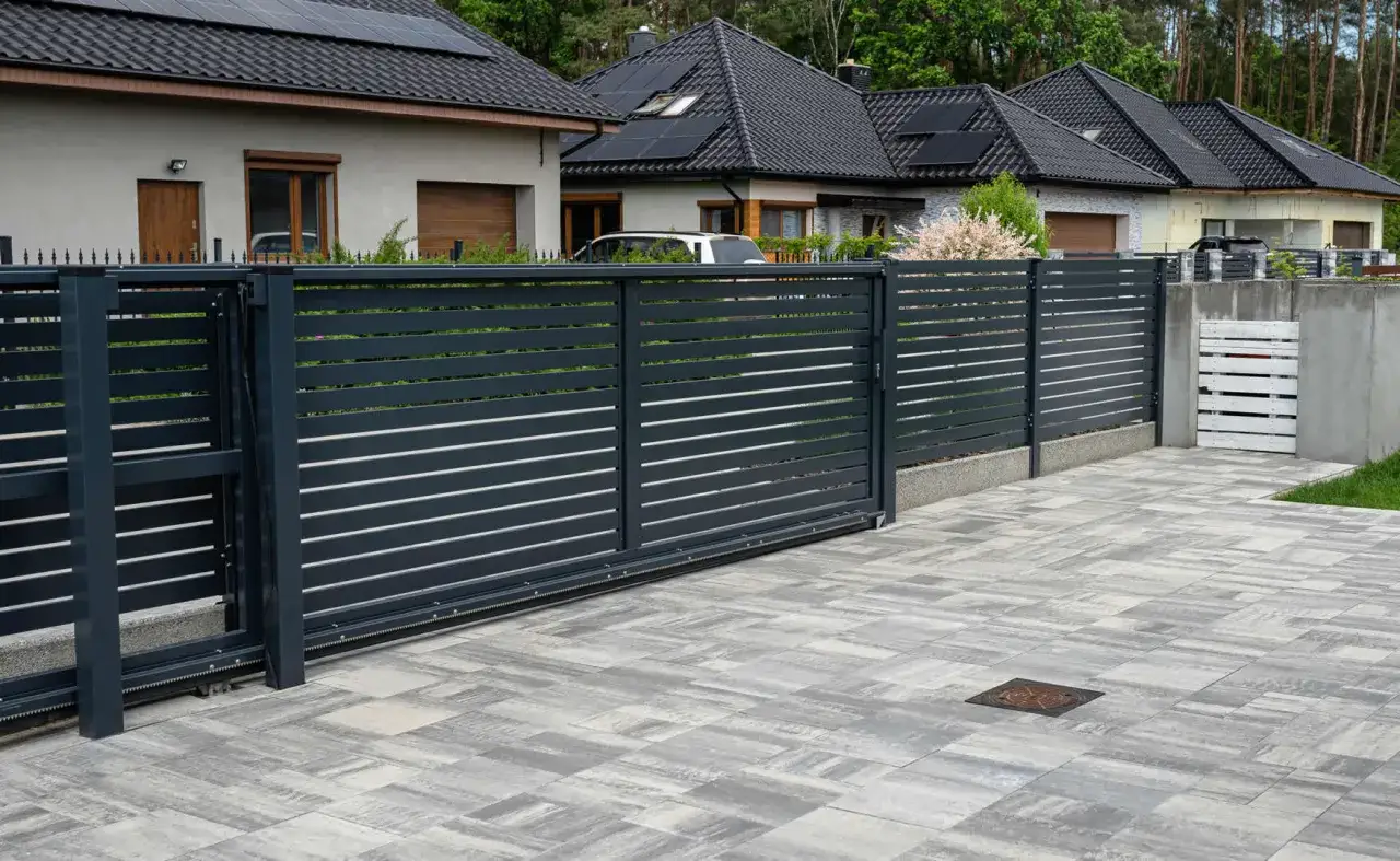 Moderne gartenzaun ideen bilder: dunkles Metalltor mit horizontalen Lamellen, gepflasterte Einfahrt, H&auml;user mit Solaranlagen im Hintergrund.
