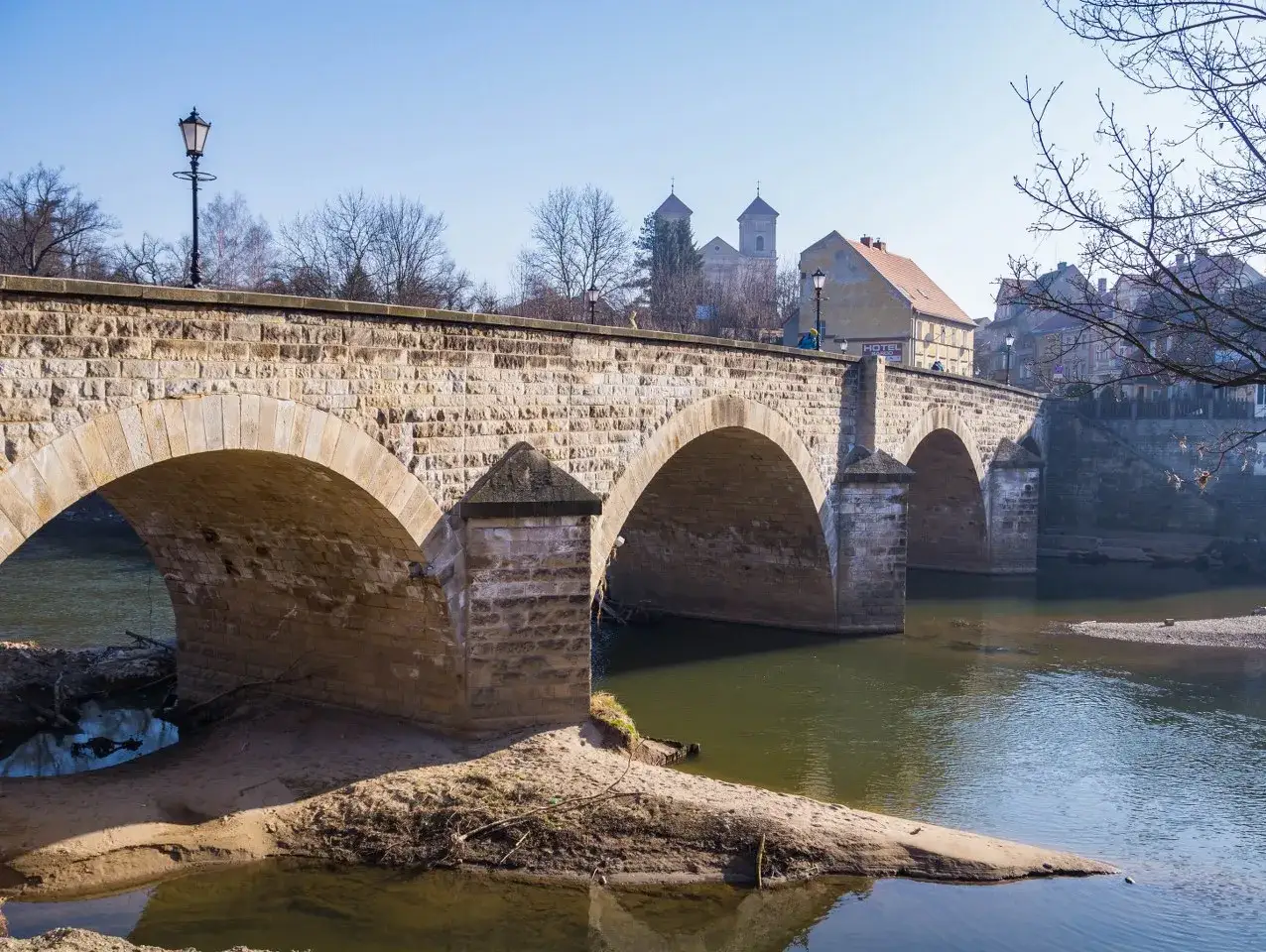 Kamienny most z arkadami nad rzeką w Bardo Śląskim. W tle kości&oacute;ł i zabudowania miejskie.