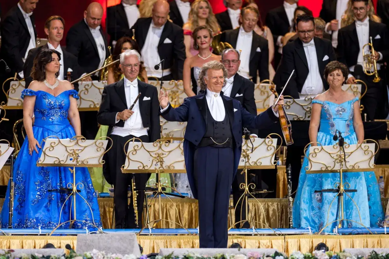 André Rieu w Polsce. Dyrygent z orkiestrą i solistkami w błękitnych sukniach na scenie.