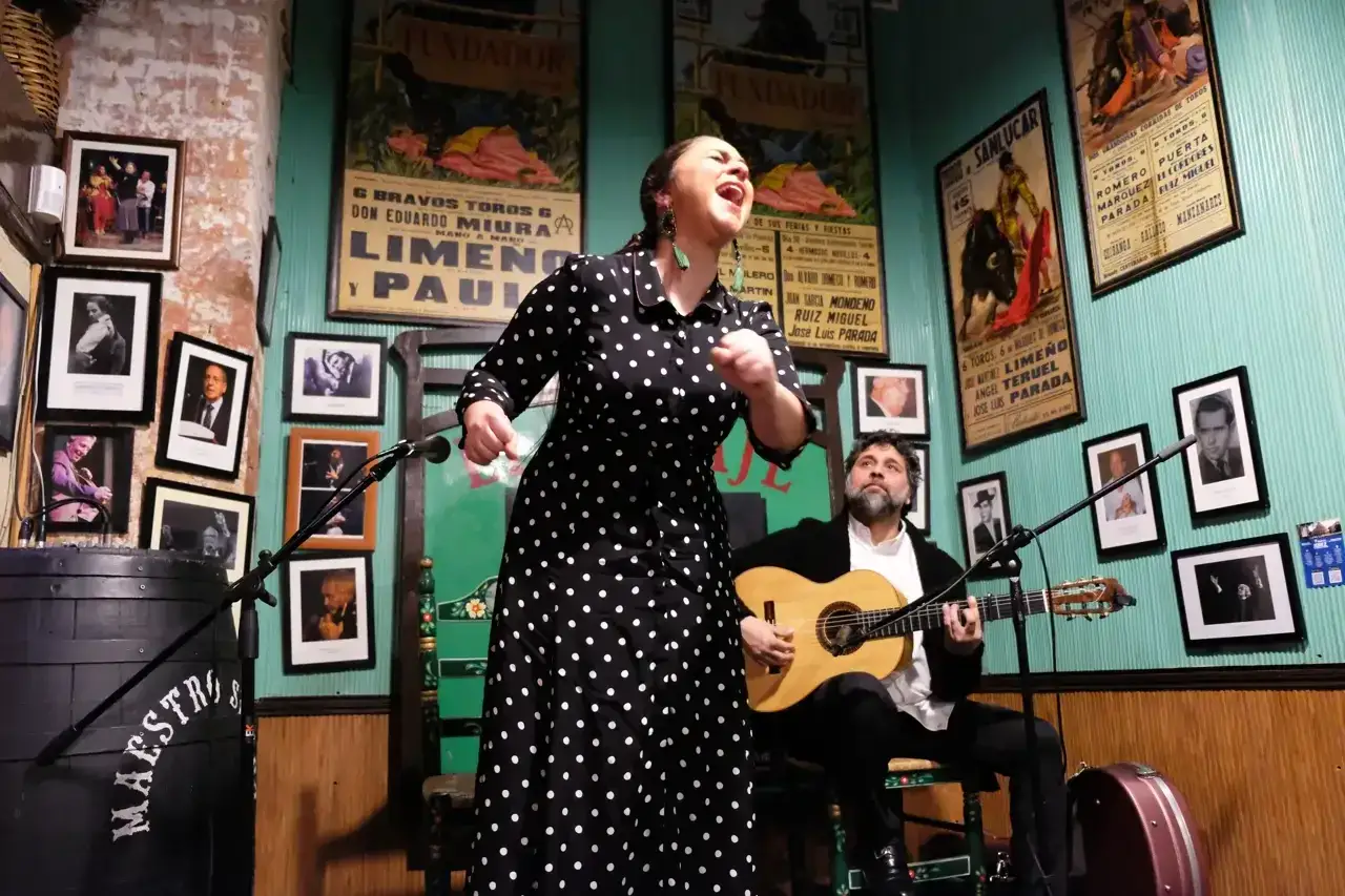 Una mujer canta con pasión, capturando las **expresiones jerezanas**, mientras un guitarrista la acompaña en un escenario íntimo.