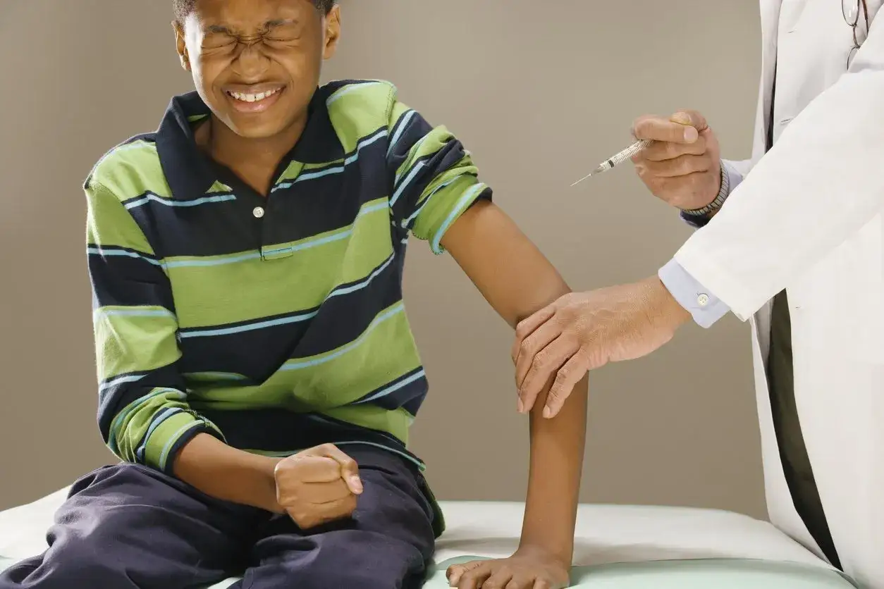 Jongen met fobie voor naalden knijpt ogen dicht terwijl arts een injectie voorbereidt.