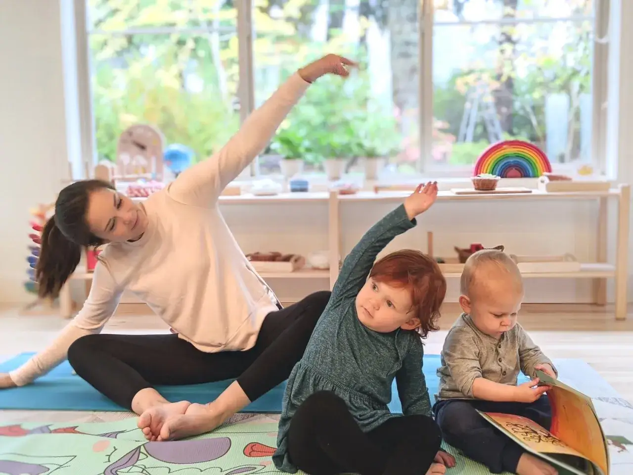 Frau macht Yoga mit Kindern unter 3 Jahren. Sie sitzen auf Matten, ein Kind liest ein Buch.