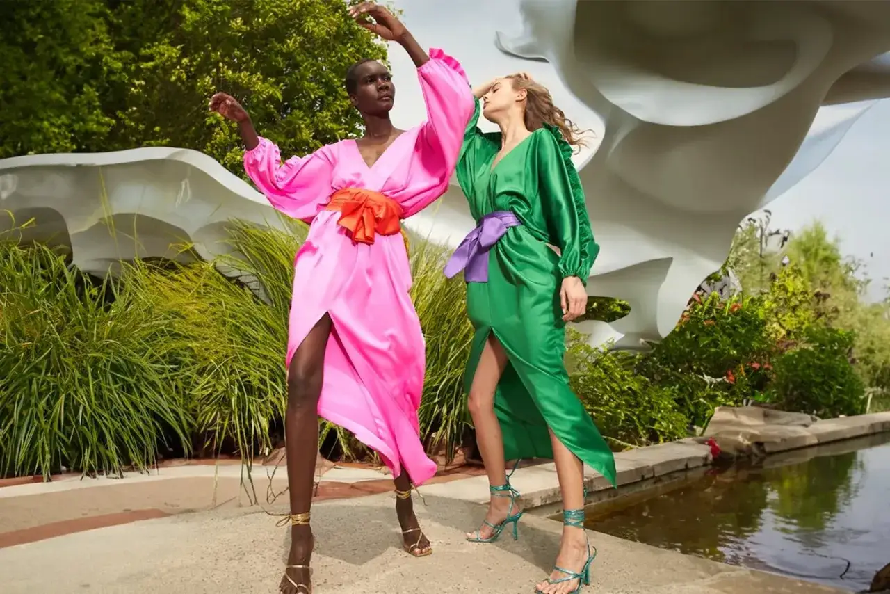 Cynthia Rowley co to za marka? Dwie kobiety w jaskrawych sukniach, różowej i zielonej, tańczą na tle nowoczesnej rzeźby i bujnej zieleni.