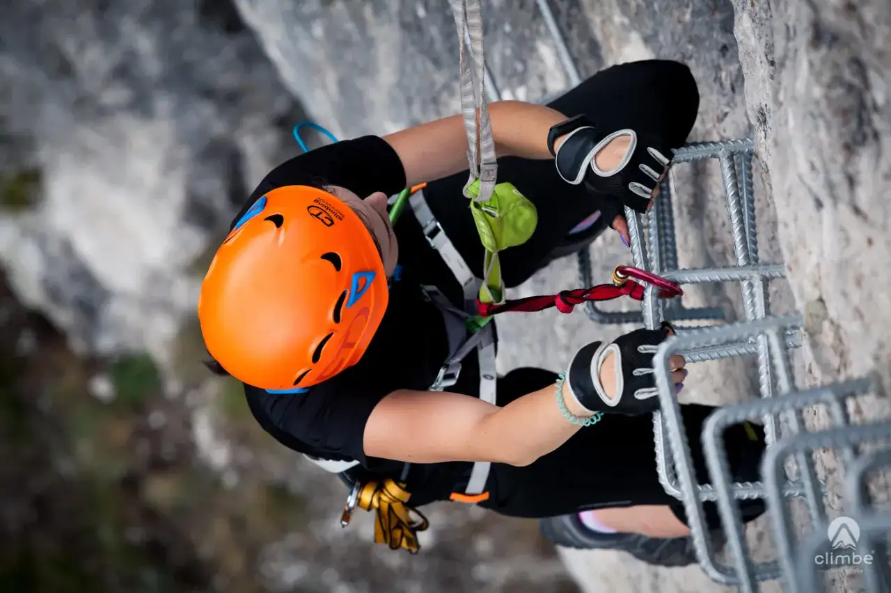 Kobieta w kasku i rękawiczkach wspina się po drabinie na via ferrata, zabezpieczona liną.