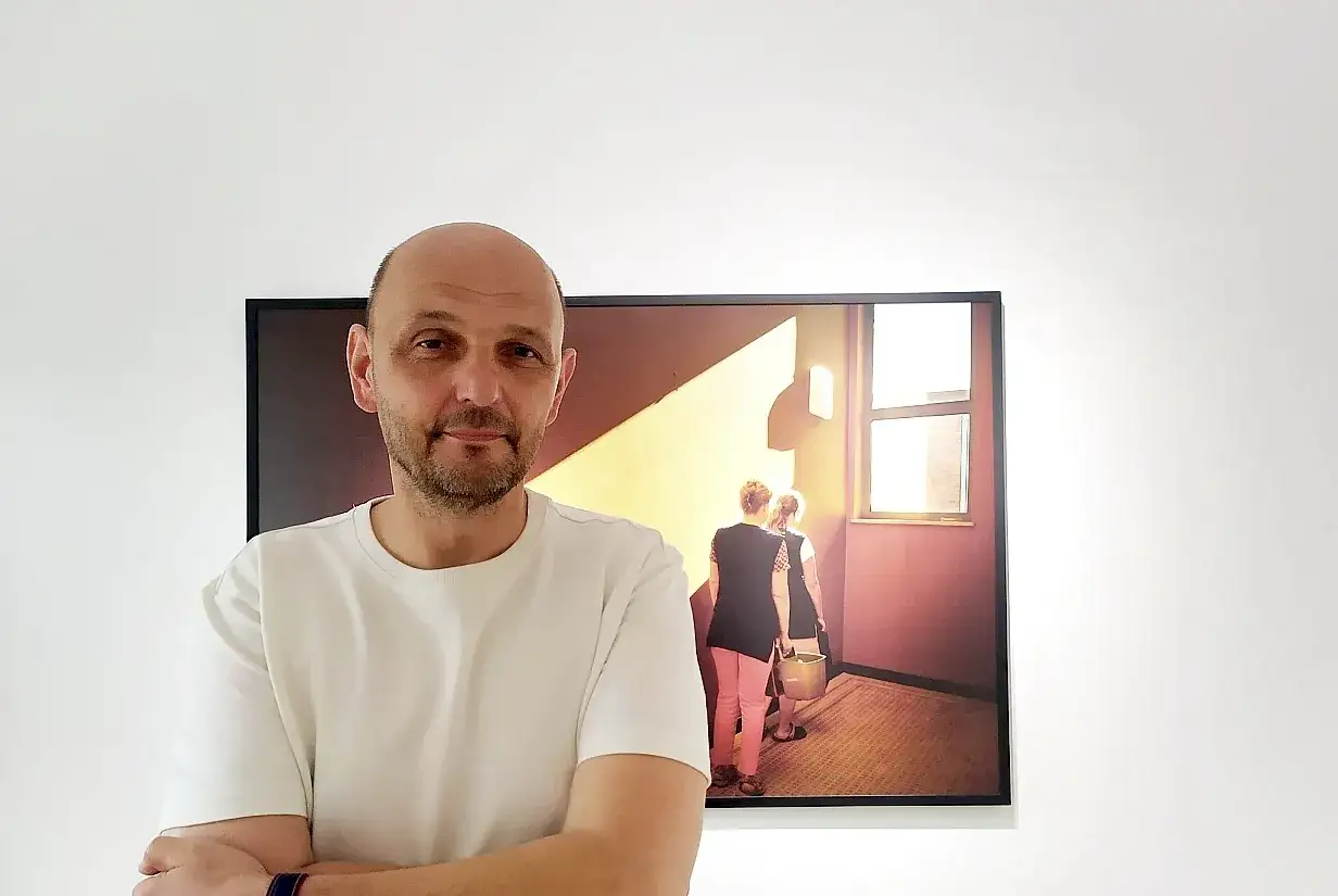 Krzysztof Gołuch obok swojego dzieła, na którym dwie kobiety idą korytarzem oświetlonym słońcem.
