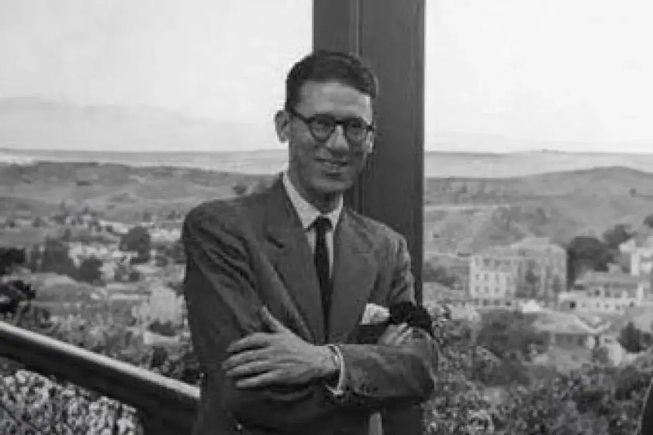 José Luis Cano, con gafas y traje, sonríe con los brazos cruzados ante un paisaje urbano y montañoso.