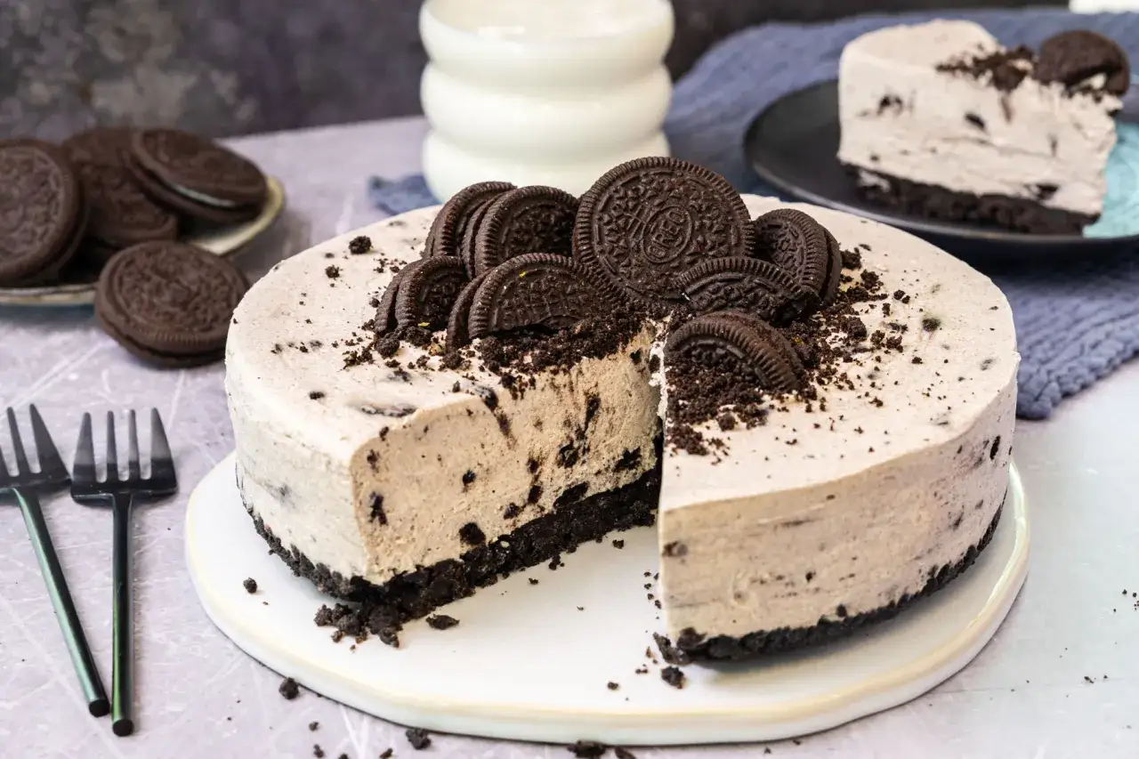 Ein k&ouml;stlicher Oreo Kuchen, verziert mit ganzen und zerbrochenen Keksen, wartet darauf, angeschnitten zu werden. Daneben liegen Gabeln und ein Glas Milch.