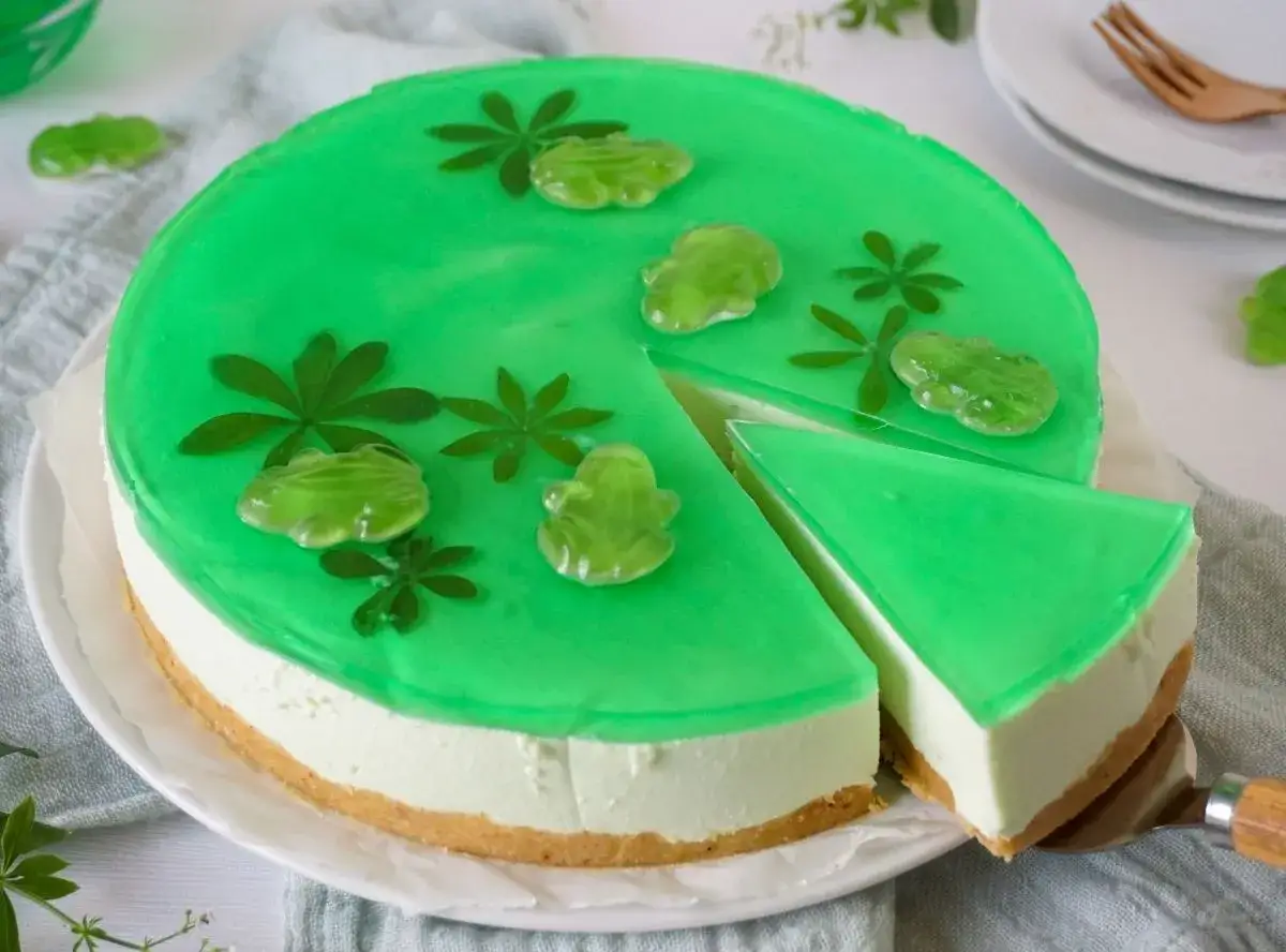 Eine gr&uuml;ne Philadelphia Torte mit Waldmeister-Gelee, verziert mit Gummifr&ouml;schen und Bl&auml;ttern. Ein St&uuml;ck wird gerade herausgeschnitten.
