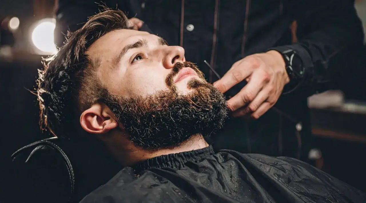 Stylista precyzyjnie przycina brody męskie krótkie. Włosy na twarzy mężczyzny są ciemne i gęste.