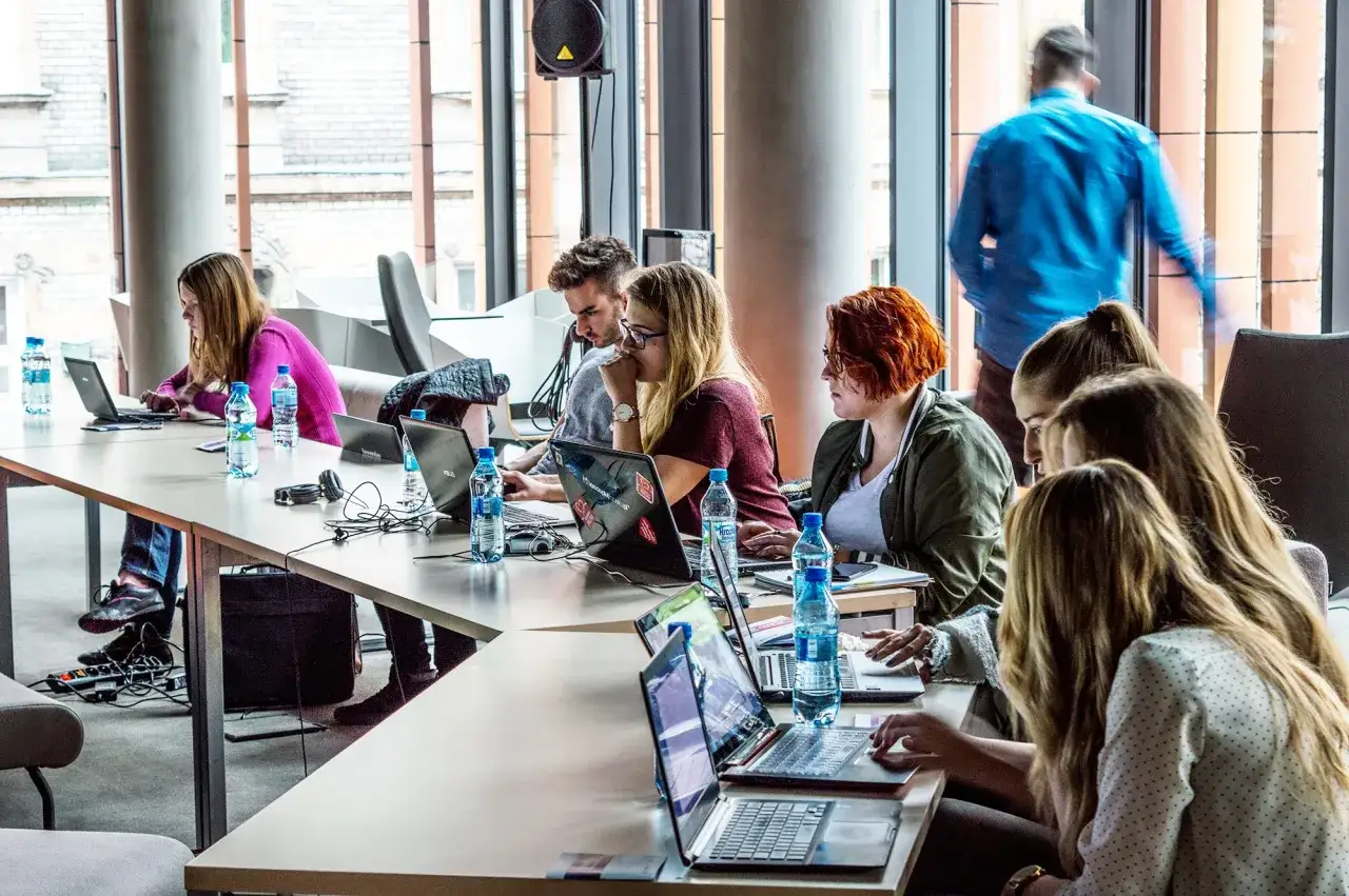 Grupa osób pracuje przy laptopach, dyskutując o tym, czy bootcamp czy warto. W tle widać mężczyznę w niebieskiej koszuli.
