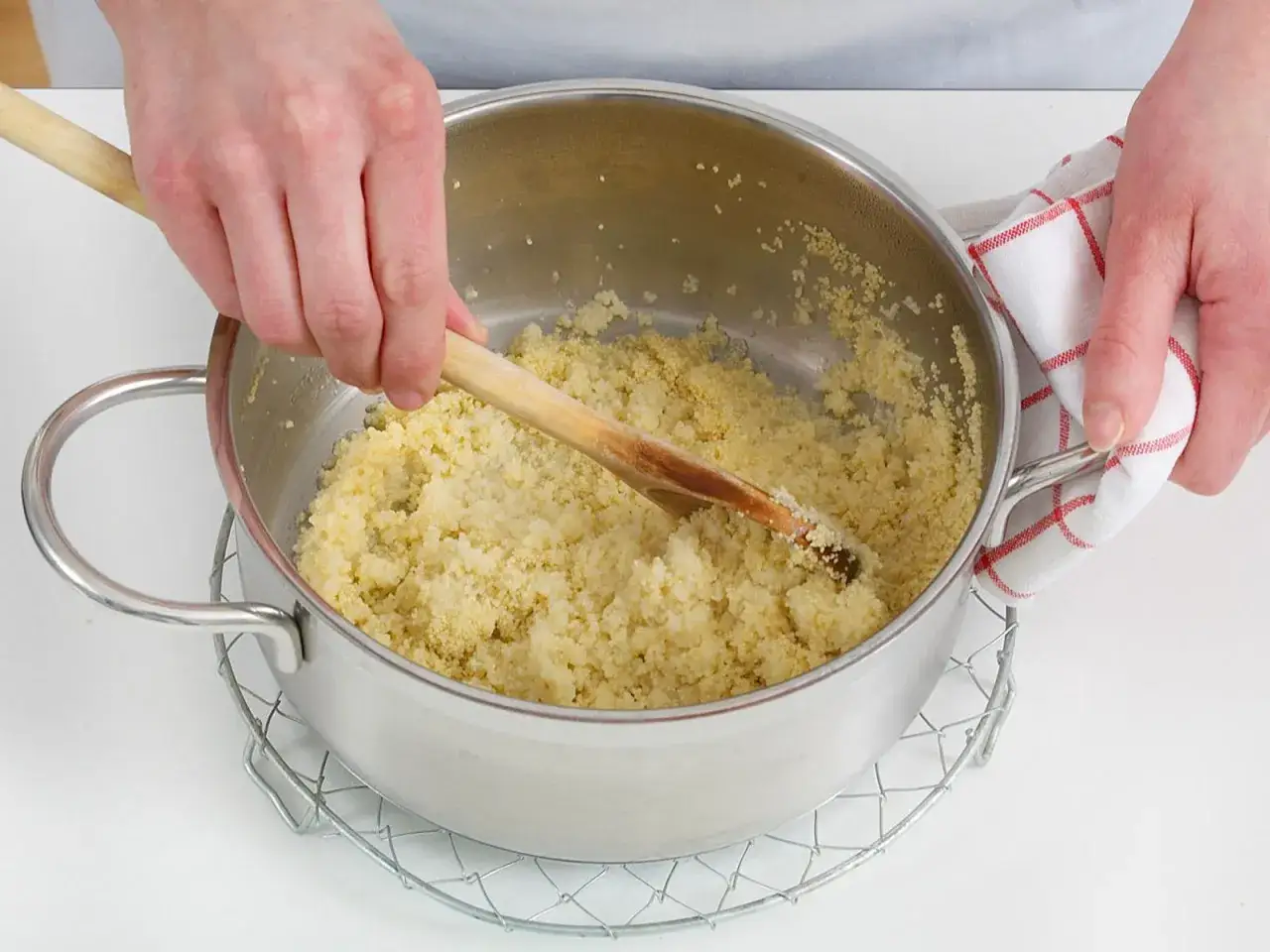 H&auml;nde r&uuml;hren mit einem Holzl&ouml;ffel in einem Topf, um Couscous zuzubereiten. Ein K&uuml;chentuch h&auml;lt den Topf.