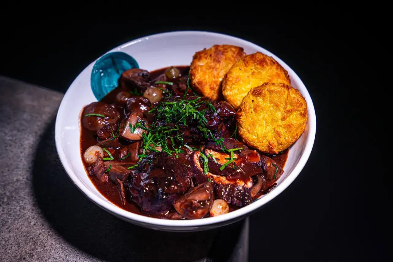Ein Teller mit Coq au Vin Rezept Original: zartes Hähnchen in Rotwein geschmort, mit Pilzen und Perlzwiebeln, garniert mit frischen Kräutern und serviert mit knusprigen Kartoffelpuffern.