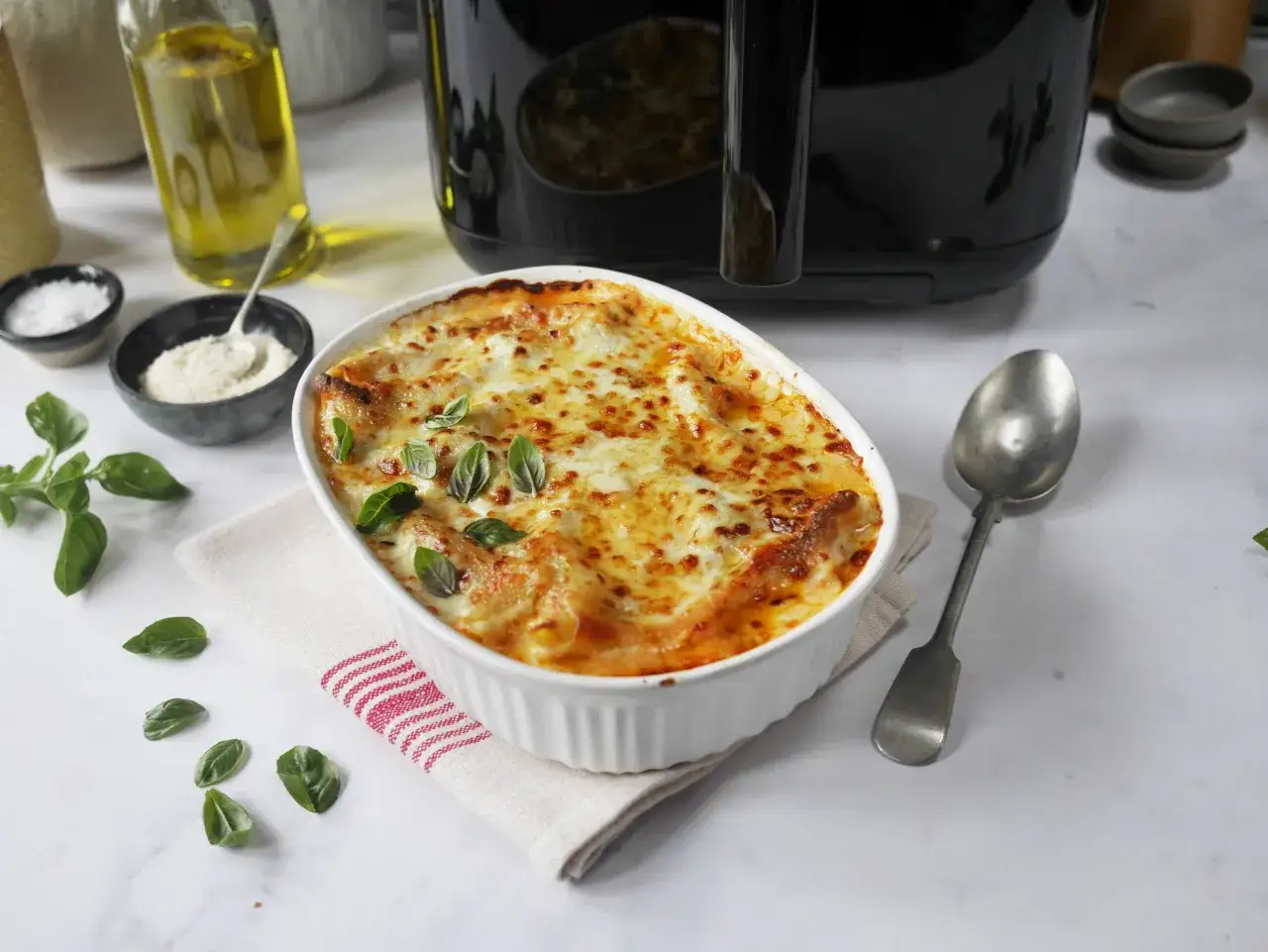 En gyllene lasagne i airfryer, toppad med smält ost och färsk basilika. Serveras i en vit ugnsform med en sked bredvid.