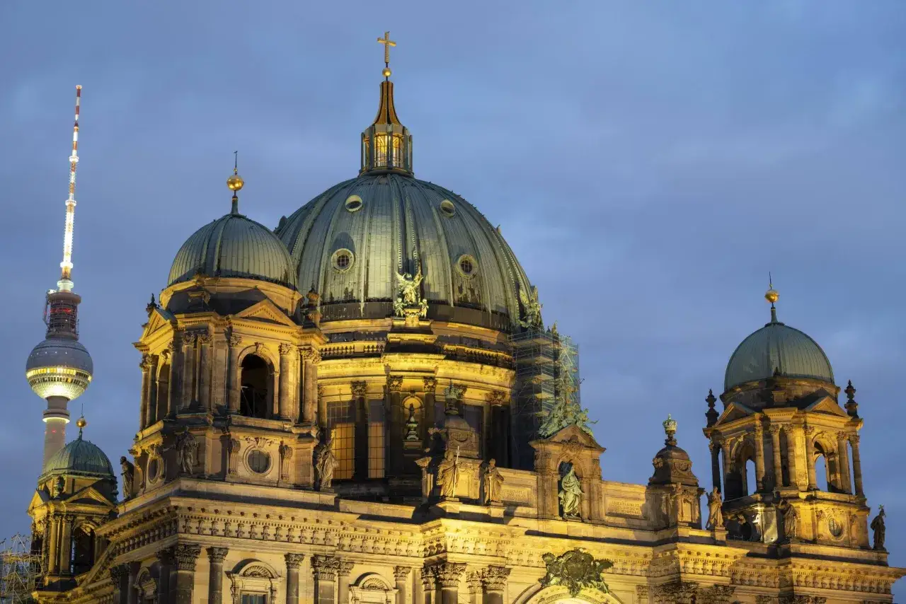 Prachtvolle Kuppeln des Berliner Doms im Abendlicht, mit dem Fernsehturm im Hintergrund. Ein Wahrzeichen der Kirchen in Berlin.
