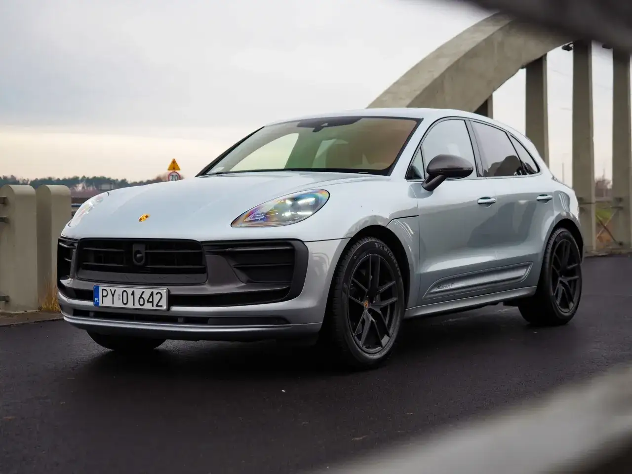 Srebrny Porsche Macan T, jeden z najnowszych modeli, prezentuje się imponująco na tle mostu.