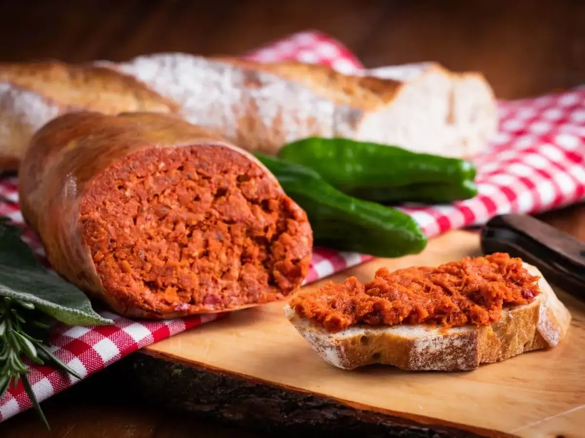 Ein Stück Brot mit scharfer, roter Nduja, einer italienischen Streichwurst. Daneben ein Anschnitt der Wurst, grüne Paprika und Baguette.