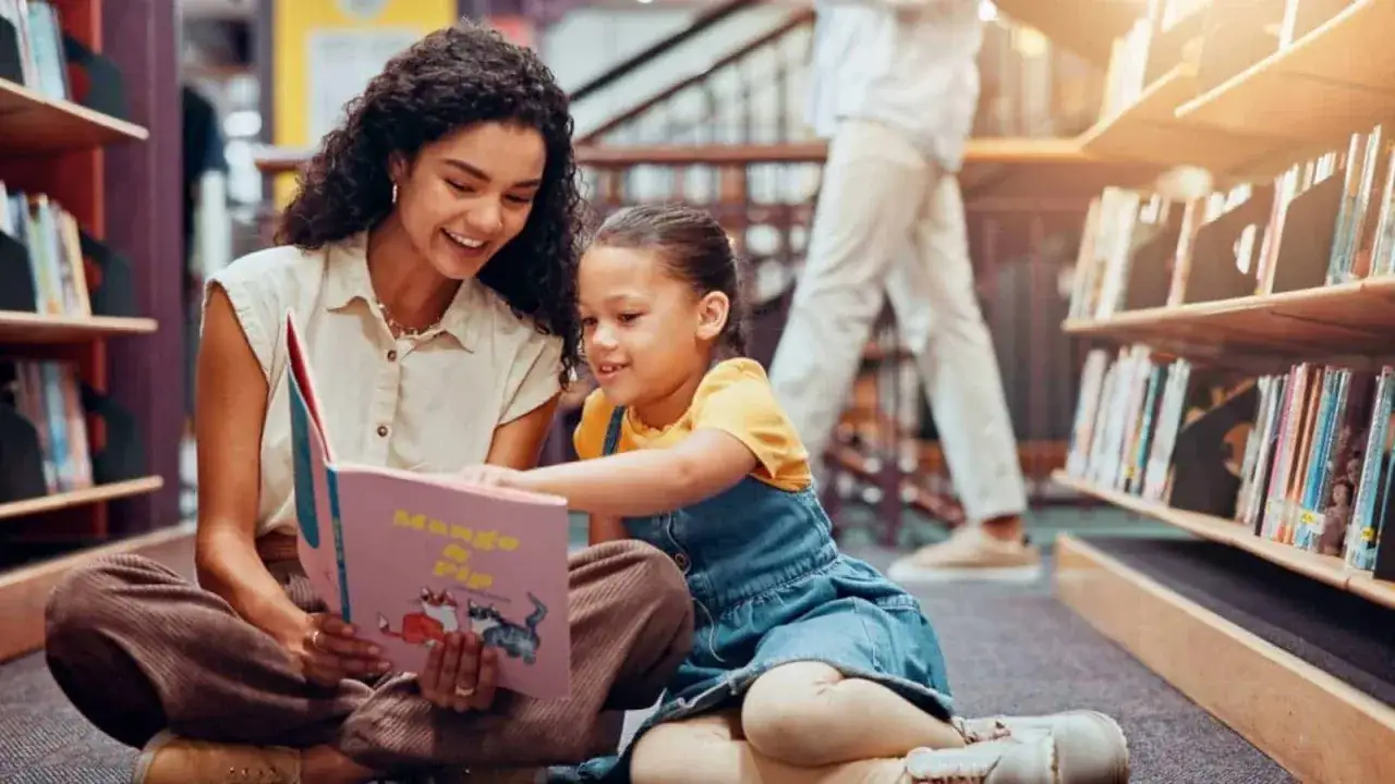 Eine Frau und ein Kind sitzen auf dem Boden einer Bibliothek und lesen ein Buch. Sie sind umgeben von Bücherregalen, was auf eine Atmosphäre von Bewegung und Lernen hindeutet.