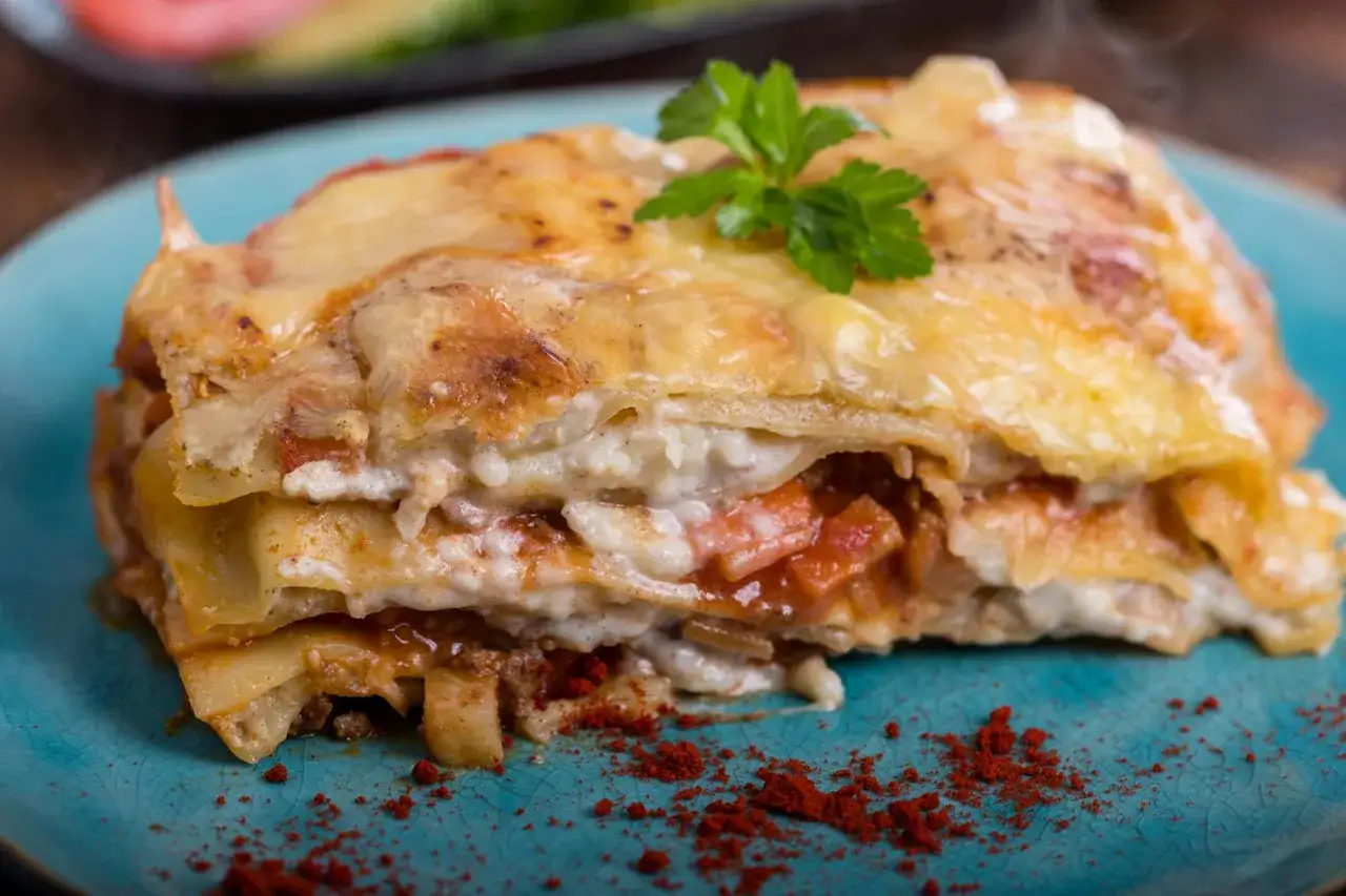 Ein St&uuml;ck Lasagne mit Schmand, &uuml;berbacken mit K&auml;se und garniert mit Petersilie, auf einem blauen Teller.
