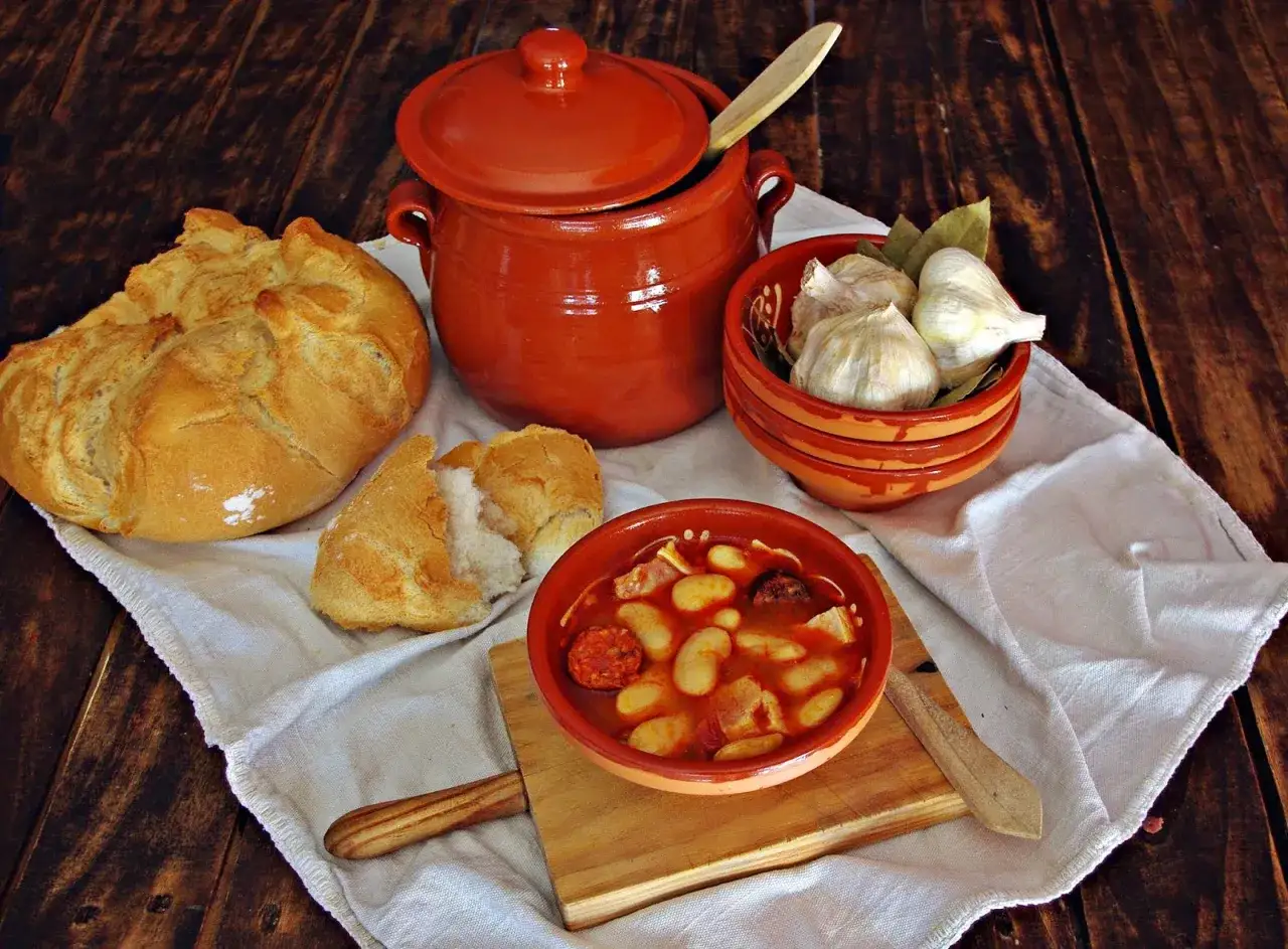 Un plato humeante de judiones de la granja, con pan r&uacute;stico, ajos y una olla de barro.