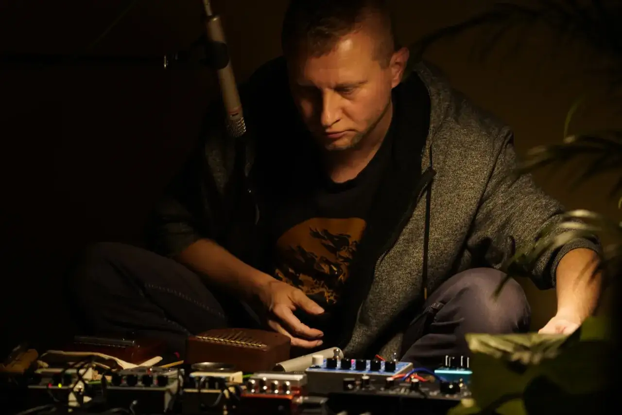 Rafał Iwański, muzyk, siedzi przy mikrofonie, otoczony efektami gitarowymi i kalimbą, tworząc dźwięki.