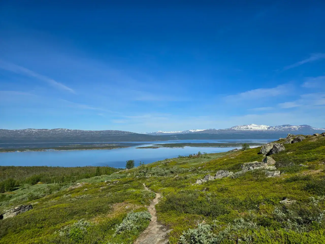 Vacker vy &ouml;ver en sj&ouml; och sn&ouml;kl&auml;dda berg i Padjelanta nationalpark. En stig slingrar sig genom gr&ouml;nskande fj&auml;llhed.