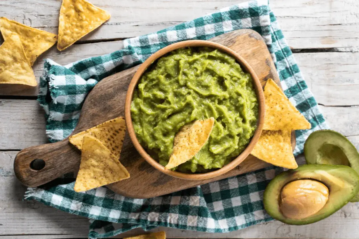 Miseczka z najpyszniejszym guacamole, otoczona chrupiącymi nachosami i świeżymi awokado, na drewnianej desce.