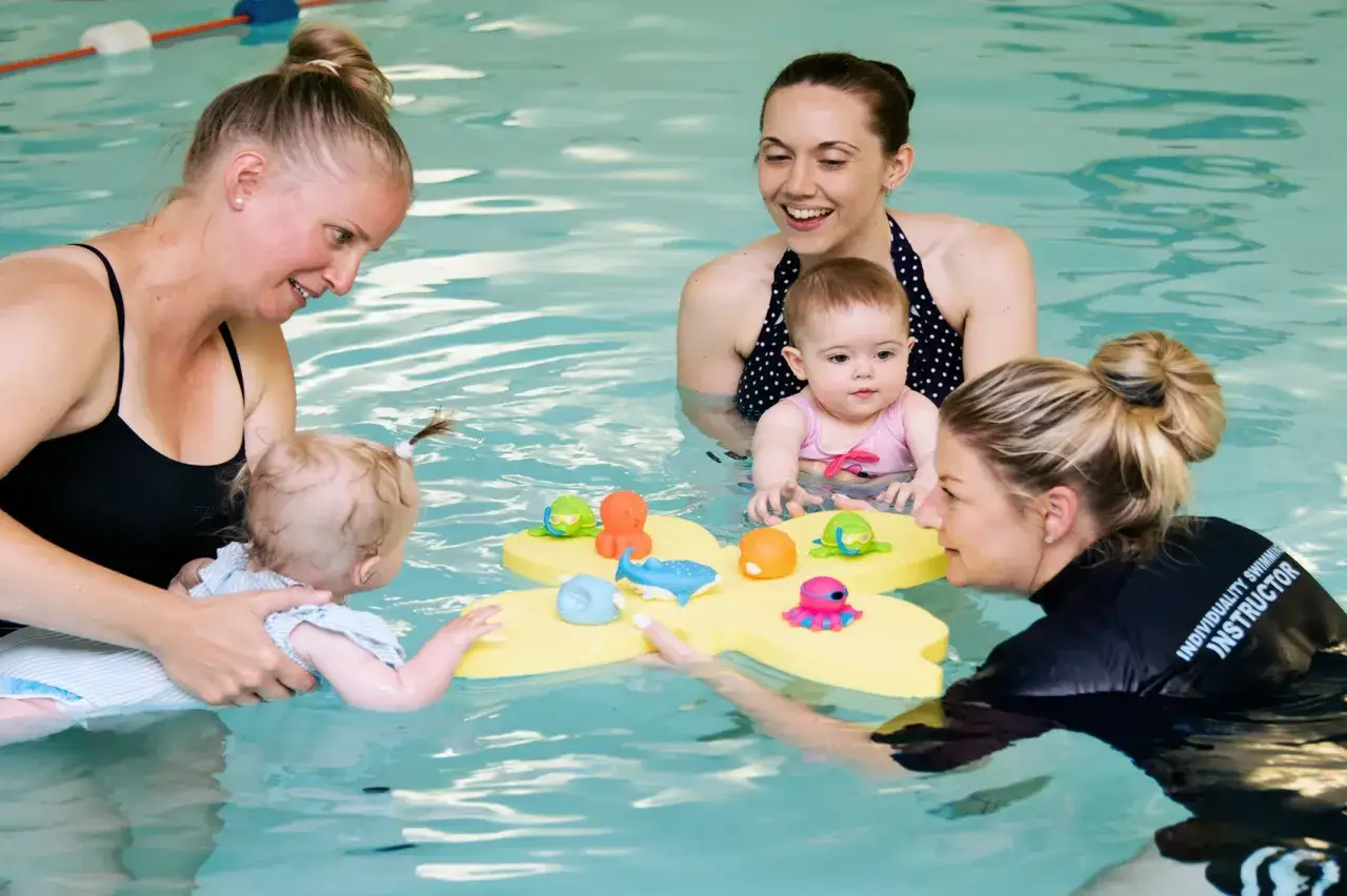 Mothers and instructors engage toddlers in a fun toddler swim school class, playing with colorful bath toys in a pool.