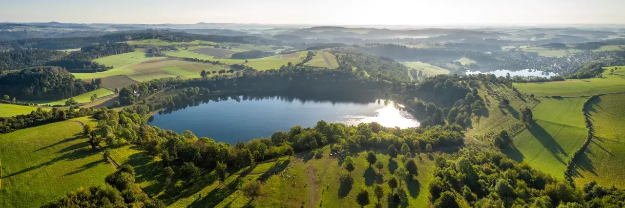 Idyllische Landschaft mit einem Maar, umgeben von grünen Hügeln und Feldern. Ein perfekter Ort für einen rundweg um die drei dauner maare.
