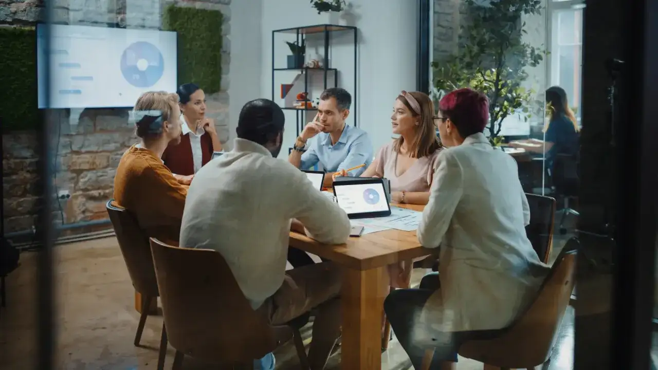 Team members engage in an effective communication presentation, discussing strategy on laptops and tablets around a wooden table.